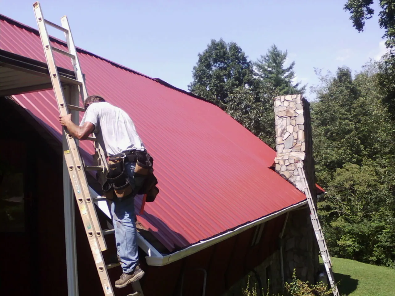 Roofer on a ladder beside a red metal roof and stone chimney — Soffit & Fascia Repair by Roofing Brothers in Clinton Corners